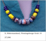 auf Stoff fotografiert:Glasperlenkette aus weißen, taubengrauen und blauen Perlen;eine große schwarze mit gelben Flecken und Achterschleife, eine türkise mit rot-weißer Fasdenauflage