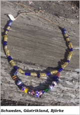 Eine Glasperlenkette mit vielen goldenen und einigen blauen Perlen, an derem unteren Ende eine zweite Kette aus verschiedenen bunten Perlen eingehängt ist.